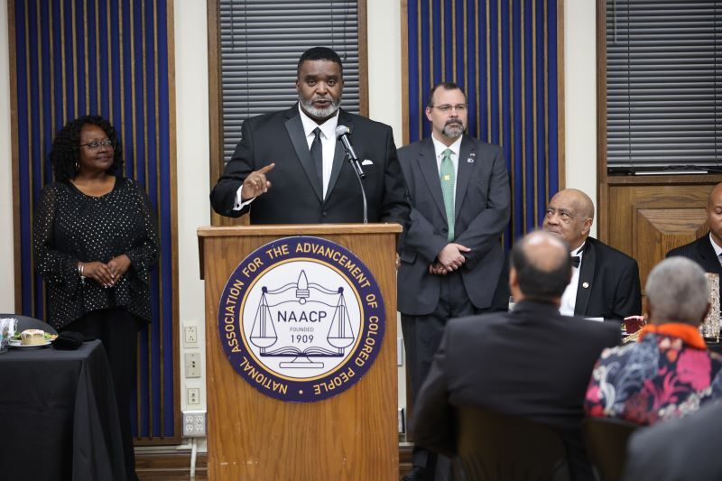 A man standing at a podium talking to an audience with a man and woman on either side of him. 